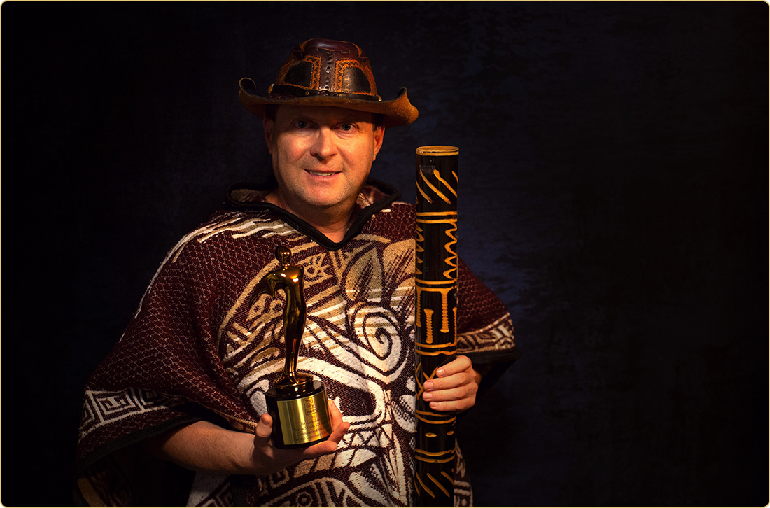 Award-winning filmmaker Arek Zasowski in a tribal poncho and adventure hat, proudly holding his Gold Telly Award and rainstick—evoking cinematic adventure and global storytelling