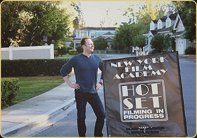 Arek Zasowski on set during NYFA filming at Universal Studios backlot Arek Zasowski stands beside a New York Film Academy “Hot Set” sign during a filming workshop on the Universal Studios Hollywood backlot, wearing a navy long-sleeve shirt and jeans