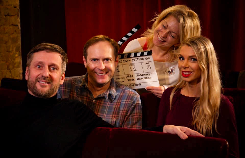 Behind-the-scenes photo from the film Let Go: Letting Go, featuring Arek Zasowski, Viola Zasowska, Paul A. Taylor, and Olga Malchevska smiling together on set, holding a clapperboard inside a theatre with red curtains.